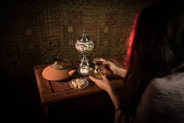 Glass of black tea in woman`s hand at Low light lounge interior with carpet. Eastern tea concept. Eastern snacks on vintage wooden surface. Selective focus
