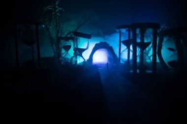 Zaman kavramı. Kum saatleri arasında duran, dumanlı ve karanlık arka planda ışıkları olan bir adamın silueti. Gerçeküstü dekore edilmiş resim