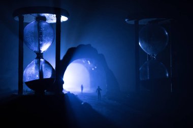 Zaman kavramı. Kum saatleri arasında duran, dumanlı ve karanlık arka planda ışıkları olan bir adamın silueti. Gerçeküstü dekore edilmiş resim