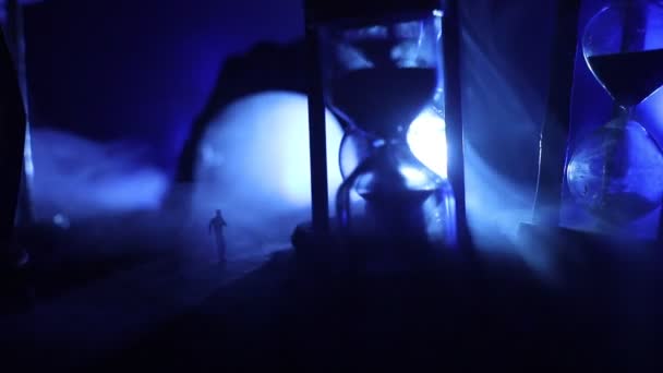 Zeitkonzept. Sand fließt durch die Glühbirnen einer Sanduhr, die die vergehende Zeit misst, während sie bis zu einem Stichtag herunterzählt. Silhouette einer Sanduhr in Rauch auf dunklem Hintergrund. mit Blumen — Stockvideo