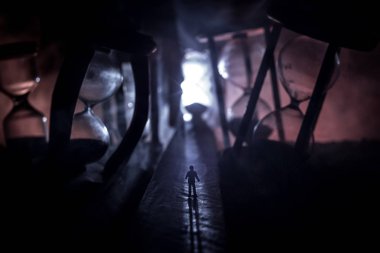 Zaman kavramı. Kum saatleri arasında duran, dumanlı ve karanlık arka planda ışıkları olan bir adamın silueti. Gerçeküstü dekore edilmiş resim