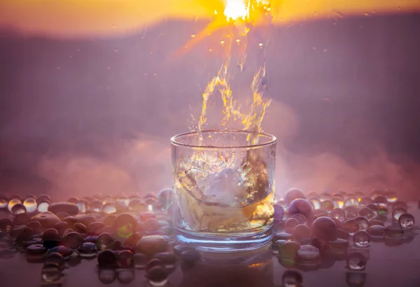 Ein Glas Whisky mit Eis und goldenem Sonnenlicht. Außenaufnahme von Whisky mit Spritzer auf dem Hintergrund des Sonnenuntergangs. — Stockfoto