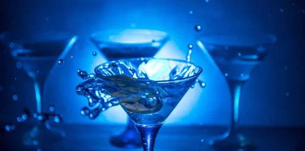 Verre à cocktail Martini à la main éclaboussant sur fond fumé foncé ou cocktail coloré en verre avec éclaboussures et olives. Soirées animées. Lumière mixte. Concentration sélective — Photo