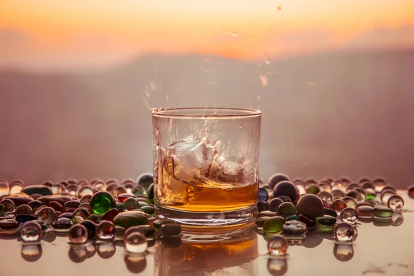 Un bicchiere di whisky con ghiaccio e luce del sole dorata. Colpo di whisky all'aperto con spruzzata sullo sfondo del tramonto . — Foto Stock