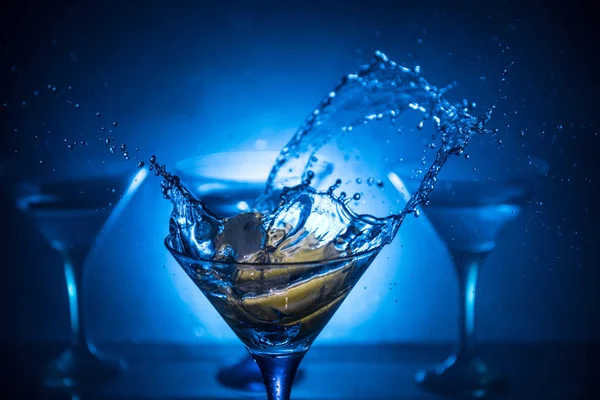 Copa de cóctel Martini en mano salpicando sobre fondo ahumado de tono oscuro o cóctel colorido en vaso con salpicaduras y aceitunas. Entretenimiento del club. Luz mixta. Enfoque selectivo —  Fotos de Stock