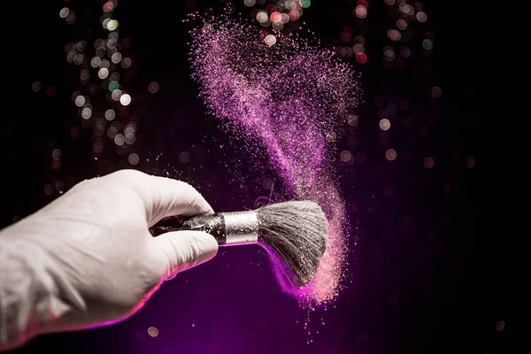 Cepillo de maquillaje en mano con polvo cosmético sobre fondo oscuro con luz y humo. Salpicadura de polvo en la oscuridad — Foto de Stock