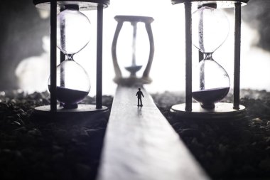 Zaman kavramı. Kum saatleri arasında duran, dumanlı ve karanlık arka planda ışıkları olan bir adamın silueti. Gerçeküstü dekore edilmiş resim