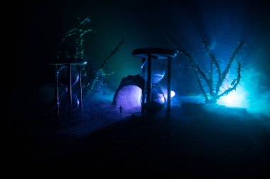 Zaman kavramı. Kum saatleri arasında duran, dumanlı ve karanlık arka planda ışıkları olan bir adamın silueti. Gerçeküstü dekore edilmiş resim