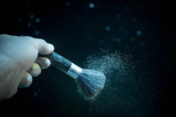 Makeup brush in hand with cosmetic powder on dark background with light and smoke. Powder splash on dark. Selective focus