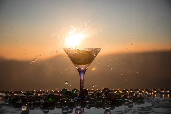 Martini cocktail glass splashing on foggy sunset background or colorful cocktail in glass with splashes and lemon. Outdoor open air party entertainment. Selective focus