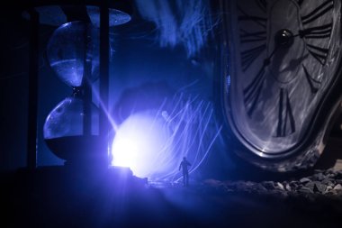 Zaman kavramı. Kum saatleri arasında duran, dumanlı ve karanlık arka planda ışıkları olan bir adamın silueti. Gerçeküstü dekore edilmiş resim