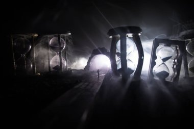 Zaman kavramı. Kum saatleri arasında duran, dumanlı ve karanlık arka planda ışıkları olan bir adamın silueti. Gerçeküstü dekore edilmiş resim