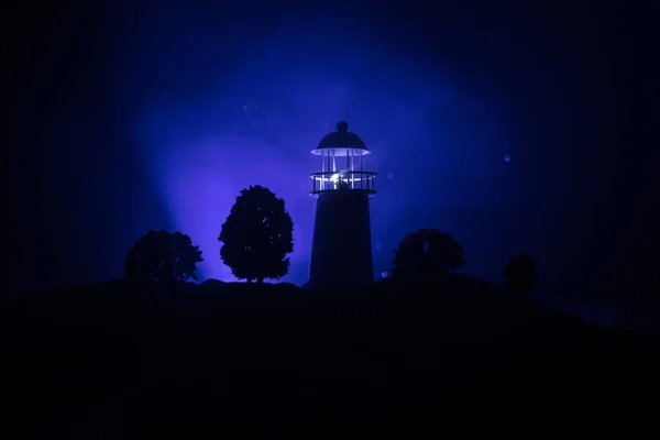 Artwork decoration. Lighthouse with light beam at night with fog. Old lighthouse standing on mountain. Table decoration. Toned background. Moonlighting. Selective focus