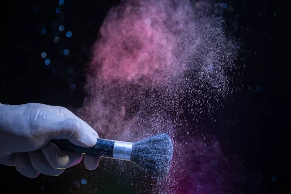 Makeup brush in hand with cosmetic powder on dark background with light and smoke. Powder splash on dark. Selective focus