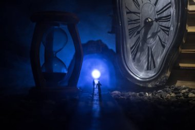 Zaman kavramı. Kum saatleri arasında duran, dumanlı ve karanlık arka planda ışıkları olan bir adamın silueti. Gerçeküstü dekore edilmiş resim