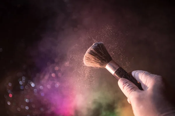 Makeup brush in hand with cosmetic powder on dark background with light and smoke. Powder splash on dark. Selective focus
