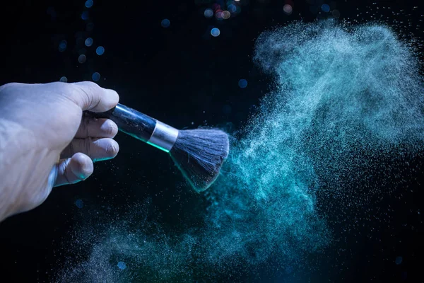 Makeup brush in hand with cosmetic powder on dark background with light and smoke. Powder splash on dark. Selective focus