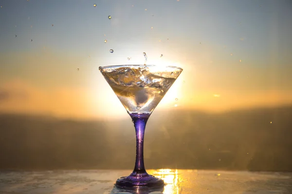 Martini cocktail glass splashing on foggy sunset background or colorful cocktail in glass with splashes and lemon. Outdoor open air party entertainment. Selective focus