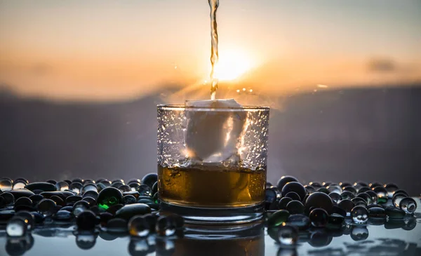 A Glass Of whiskey with ice and gold sunlight. Outdoor shot of whiskey with splash on sunset background. Selective focus