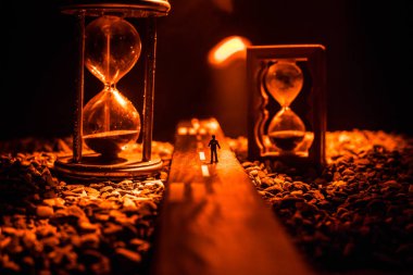 Zaman kavramı. Kum saatleri arasında duran, dumanlı ve karanlık arka planda ışıkları olan bir adamın silueti. Gerçeküstü dekore edilmiş resim