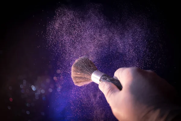 Cepillo de maquillaje en mano con polvo cosmético sobre fondo oscuro con luz y humo. Salpicadura de polvo en la oscuridad — Foto de Stock