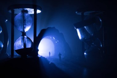 Zaman kavramı. Kum saatleri arasında duran, dumanlı ve karanlık arka planda ışıkları olan bir adamın silueti. Gerçeküstü dekore edilmiş resim