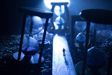 Zaman kavramı. Kum saatleri arasında duran, dumanlı ve karanlık arka planda ışıkları olan bir adamın silueti. Gerçeküstü dekore edilmiş resim