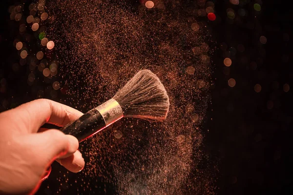 Cepillo de maquillaje en mano con polvo cosmético sobre fondo oscuro con luz y humo. Salpicadura de polvo en la oscuridad — Foto de Stock