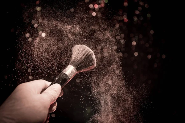 Cepillo de maquillaje en mano con polvo cosmético sobre fondo oscuro con luz y humo. Salpicadura de polvo en la oscuridad — Foto de Stock
