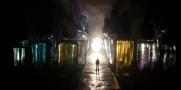 Silueta de un hombre parado en medio de la carretera en una noche brumosa con botellas de vidrio gigantes llenas de líquido de cigarrillo electrónico. Nubes de niebla coloridas con luz en el fondo . — Foto de Stock