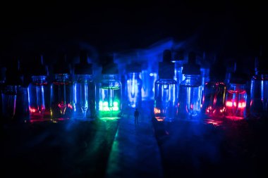 Elektronik sigara sıvısı ile dolu dev cam şişeler ile sisli bir gecede yolun ortasında duran bir adamın silueti. Arka planda ışık ile renkli sisli bulutlar.