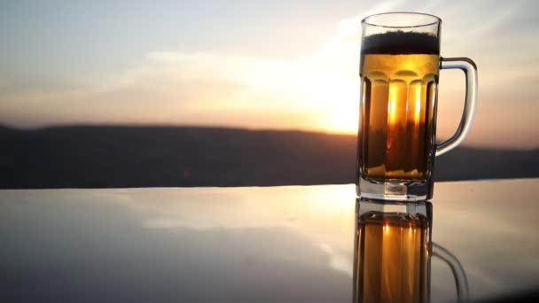 夕暮れ時のビーチでビールのグラス クーリングサマードリンクコンセプト 太陽光の背景 ソフトフォーカスのボケでドラフトビールのグラスのクローズアップ — ストック動画