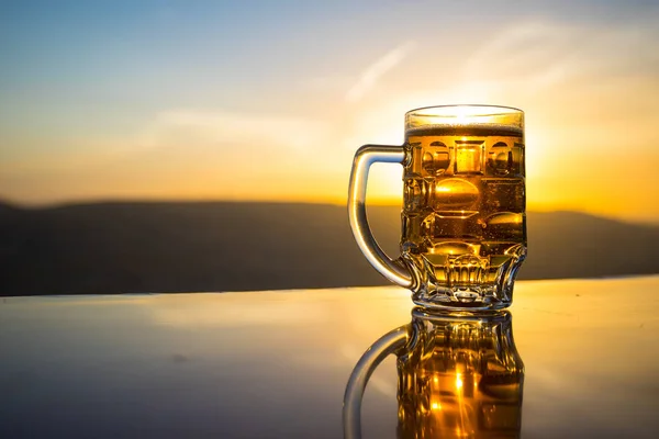 夕暮れ時のビーチでビールを飲みながら。クーリングサマードリンクコンセプト。太陽光の背景のボケでドラフトビールのグラスのクローズアップ — ストック写真