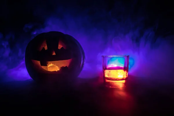 Zucca di Halloween con viso scolpito e bicchiere di whisky con ghiaccio su uno sfondo scuro tonico nebbioso con zombie. Decorato. Focus selettivo — Foto Stock