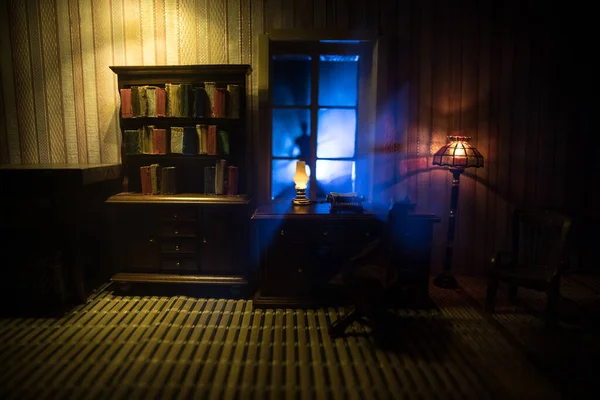 A realistic dollhouse living room with furniture and window at night. Artwork table decoration with handmade realistic dollhouse. Horror silhouette in window. Selective focus.