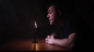 Beyaz adam haber spikeri karanlık odada mikrofonlu bir masada oturuyordu. Mikrofonlu bir sanatçı. Erkek müzisyen mikrofona şarkı söylüyor. Siyah arka planda çekilmiş. Karanlık atmosfer