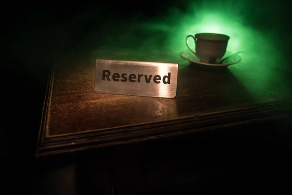 Reserved sign on the table. A tag of reservation placed on the wood table. Metal tag with reservation on dark. Reserved table in a restaurant. Creative concept with colorful lights. Selective focus