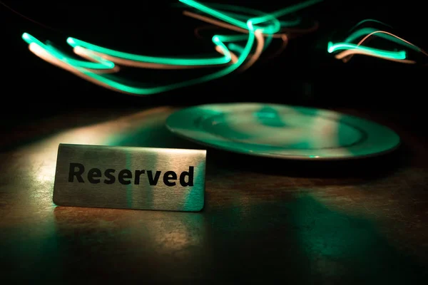 Reserved sign on the table. A tag of reservation placed on the wood table. Metal tag with reservation on dark. Reserved table in a restaurant. Creative concept with colorful lights. Selective focus