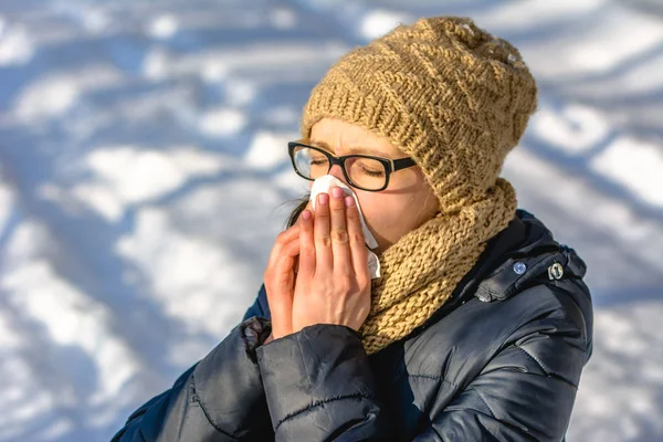 Sneezing woman blowing her nose with a tissue. Concept of people with cold and flu in winter with snow, outdoors.