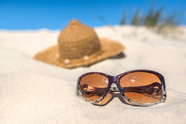 Kumsalda yaz tatili aksesuarları, kum ulağında ve mavi gökyüzünde güneş gözlüğü ve güneş şapkası ile arka plan.