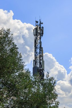 Yeşil ağaca karşı beyaz bulutlar arka plan ile mavi gökyüzünde elektronik ekipman ile telekomünikasyon kule