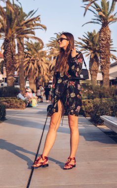 bir yaz günü açık havada poz çekici kadın. moda blogger bir çiçek mini elbise, güneş gözlüğü takıyor ve küçük bir siyah çanta tutarak. palmiye ağaçları arka plan