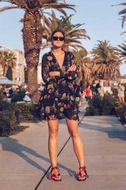 bir yaz günü açık havada poz çekici kadın. moda blogger bir çiçek mini elbise, güneş gözlüğü takıyor ve küçük bir siyah çanta tutarak. palmiye ağaçları arka plan