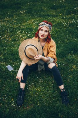 çimenlerin üzerinde oturan ve kulaklık ile poz güzel kız. Yeni müzik keşfetmek moda mutlu kadın moda Festivali giysiler dışında sıcak havalarda tadını çıkarıyor. sokak stili