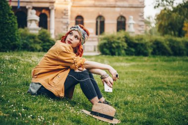 çimenlerin üzerinde oturan ve kulaklık ile poz güzel kız. Yeni müzik keşfetmek moda mutlu kadın moda Festivali giysiler dışında sıcak havalarda tadını çıkarıyor. sokak stili