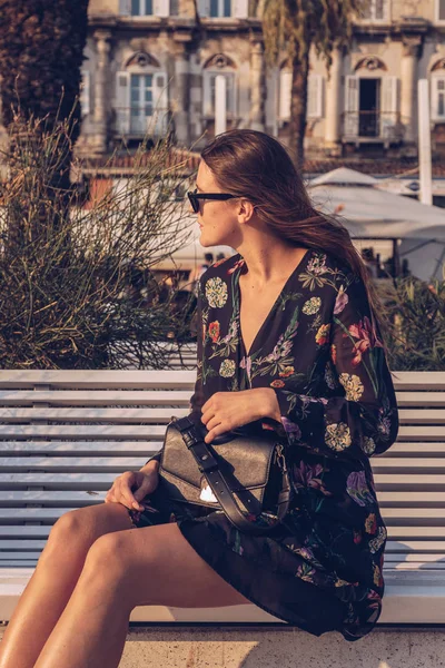 魅力的な女性は 夏の日に屋外でポーズします 花柄のミニのファッションのブロガー ドレス サングラスをかけていると 小さな黒い袋を保持しています ヤシの木の背景 — ストック写真