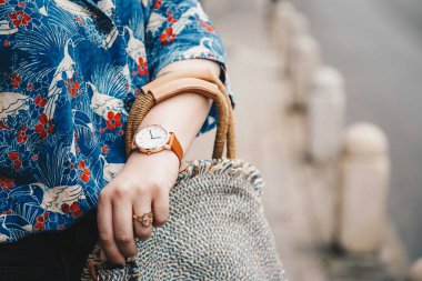 sokak tarzı moda ayrıntılar. yakın çekim, moda blogger bir yaz gömlek ve bir güzel yuvarlak hasır çanta tutan bir beyaz ve kahverengi analog kol saati takıyor. mükemmel yaz moda aksesuarları.