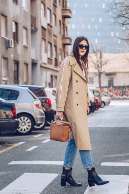 sokak tarzı bej bir palto, denim jeans, ayak bileği botları, kedi göz güneş gözlüğü ve Metal kolu kahverengi tote çanta çekici bir kadın portresi. moda kıyafet güneşli bahar günü için mükemmel
