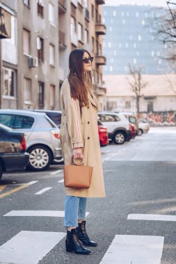 sokak tarzı bej bir palto, denim jeans, ayak bileği botları, kedi göz güneş gözlüğü ve Metal kolu kahverengi tote çanta çekici bir kadın portresi. moda kıyafet güneşli bahar günü için mükemmel
