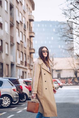 sokak tarzı bej bir palto, denim jeans, ayak bileği botları, kedi göz güneş gözlüğü ve Metal kolu kahverengi tote çanta çekici bir kadın portresi. moda kıyafet güneşli bahar günü için mükemmel